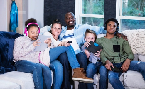 A multi-ethnic blended family with three children sitting together side by side at home on the living sofa, getting screen time.